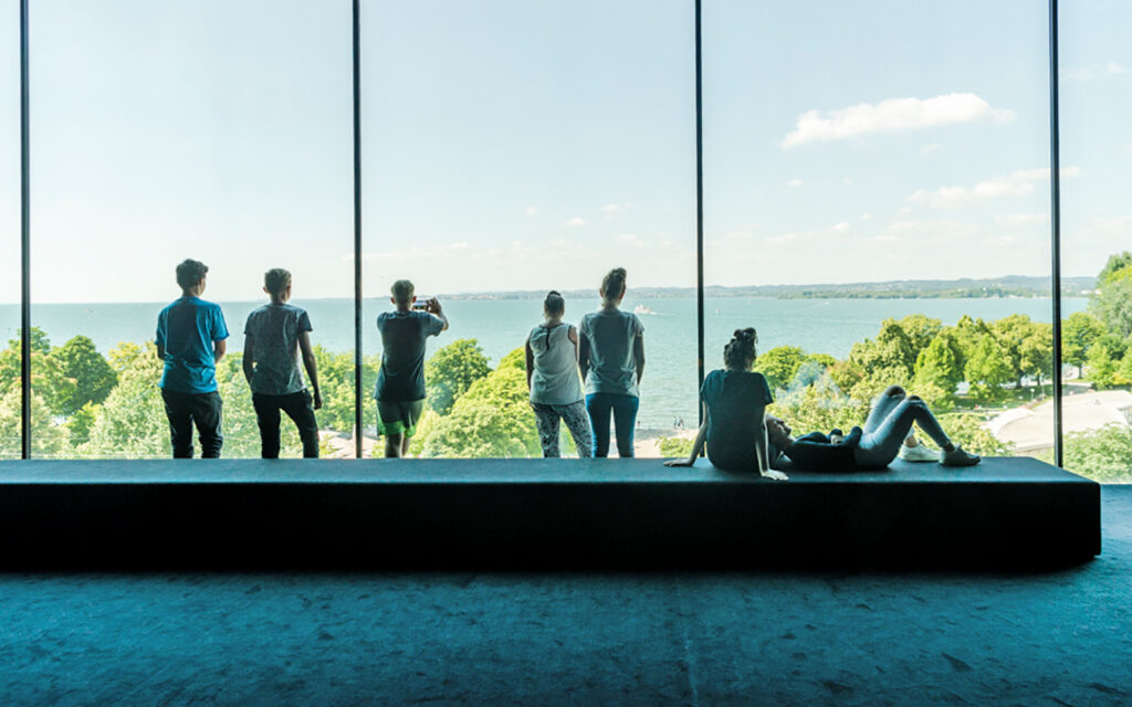 Jungen Menschen stehen im Landesmuseum Vorarlberg in einem großen Raum mit Fensterfront und schauen hinaus