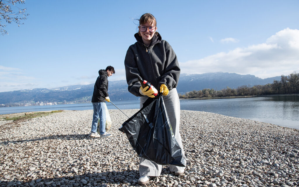 Eine junge Frau sammelt an einem Ufer Müll ein, im Hintergrund sieht man einen jungen Herrn, der auch Müll aufsammelt.