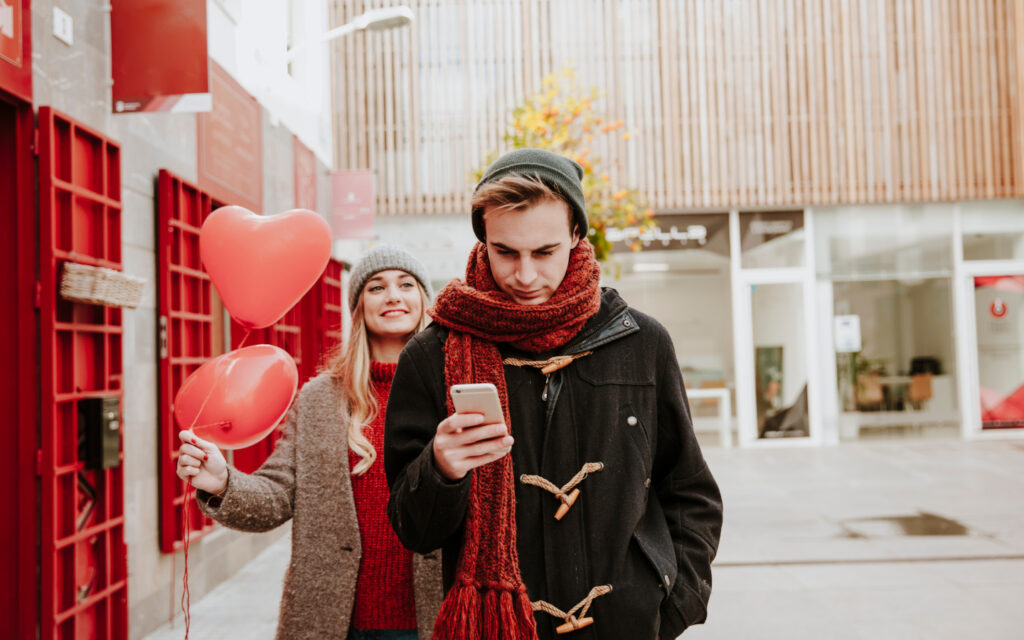 Ein Mann schaut auf sein Smartphone und läuft vor einer Frau, die Herzluftballons in der Hand hält und lächelt.