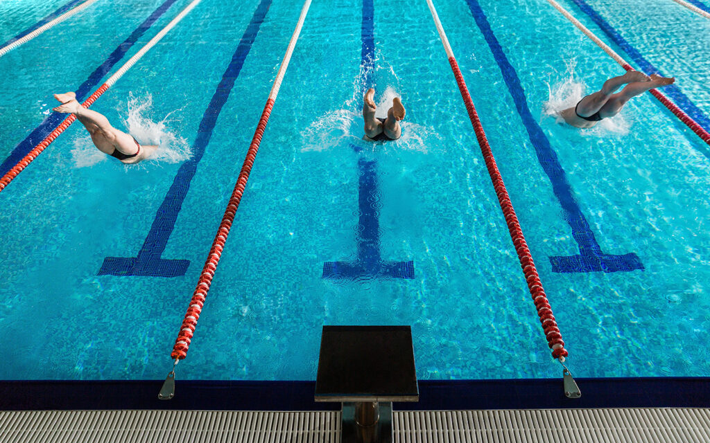 Im Hallenbad sieht man drei Menschen, die ins Becken springen