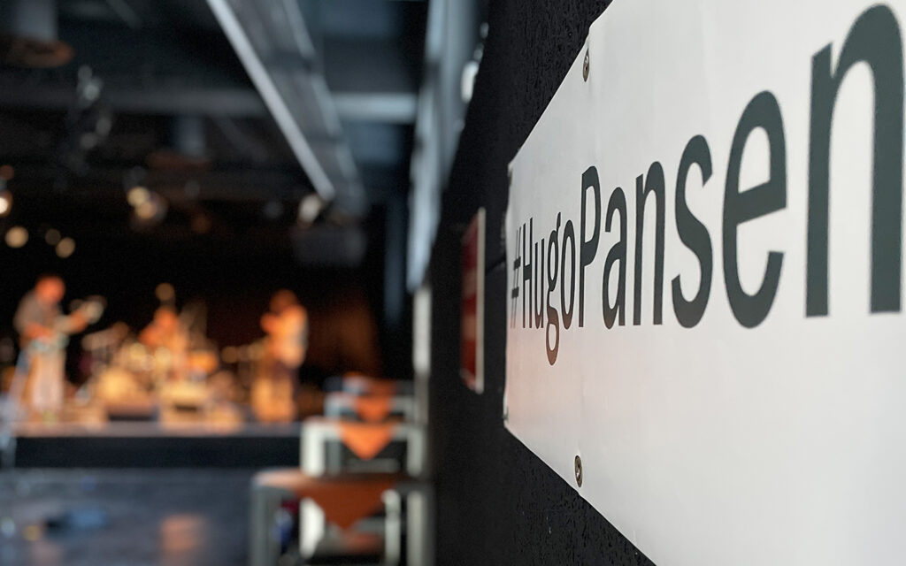 Man sieht ein Schild mit der Aufschrift "Hugo Pansen" und im Hintergrund spielen Leute auf der B&uuml;hne Musik