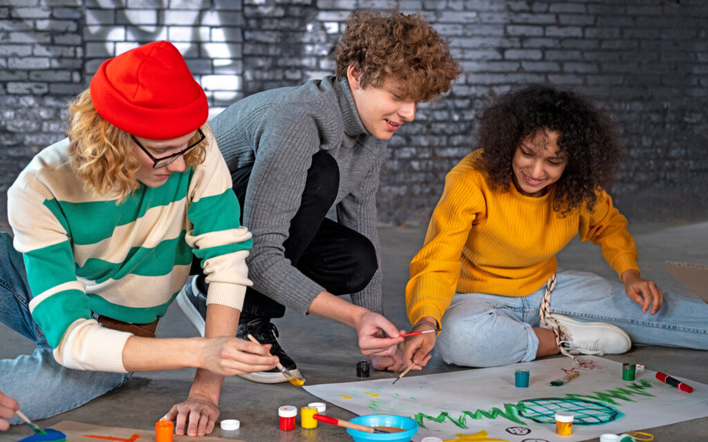 Jugendliche arbeiten gemeinsam an einem Projekt. Zwei Jungs und ein Mädchen malen am Boden ein Plakat.