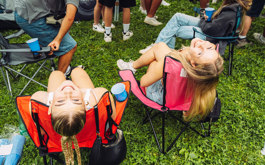 Jugendliche sitzen auf Klappsessel auf dem Campinggel&auml;nde und schauen gl&uuml;cklich in die Kamera