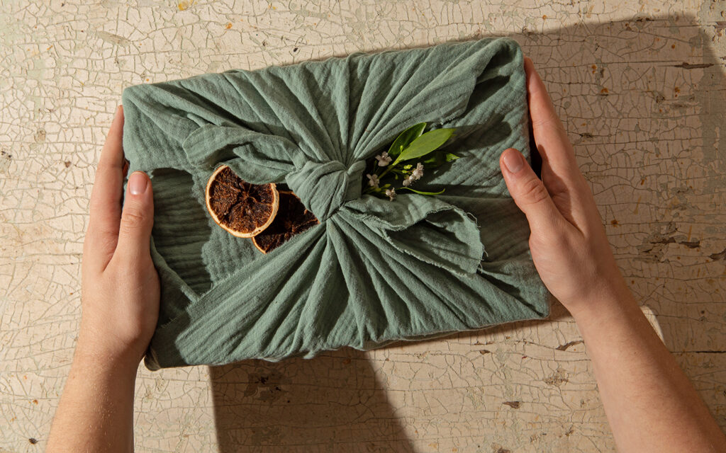Zwei H&auml;nde halten ein Geschenk in den H&auml;nden, das umweltfreundlich verpackt ist, also nur mit einem gr&uuml;nen Tuch