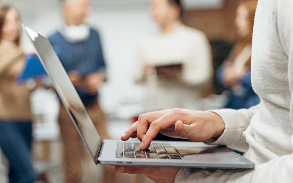 Junger Mann tippt auf seinen Laptop, im Hintergrund sind andere Menschen unscharf zu sehen