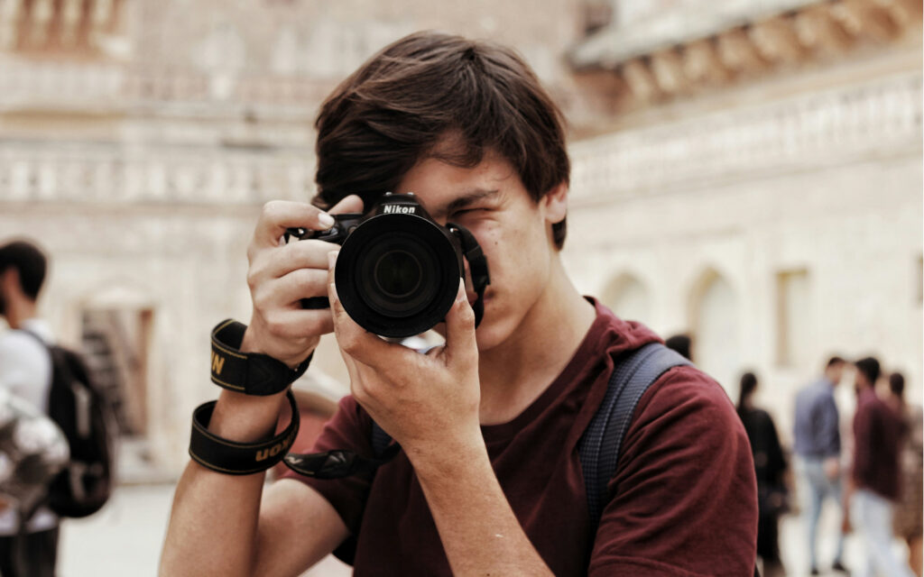 Ein junger Mann im roten T-Shirt hat eine Kamera vor dem Gesicht und fotografiert.