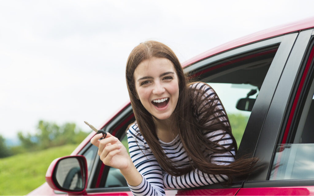 Eine Junge Frau lacht aus einem roten Auto und hält einen Schlüssel in der Hand.