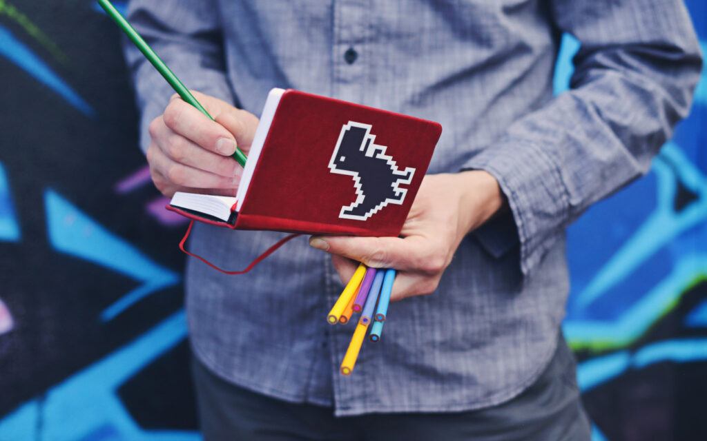 Eine Person notiert etwas in ein rotes Notizbuch mit einem Dinosaurier drauf.