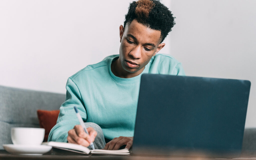 Junger Mann macht vor seinem Laptop Notizen in einen Schreibblock
