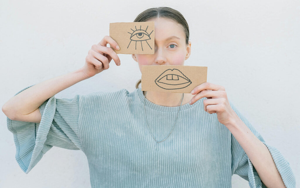 Eine junge Frau hält sich zwei Postkarten vor das Gesicht. Auf einer Postkarte ist ein Mund aufgezeichnet, auf der anderen ein Auge.