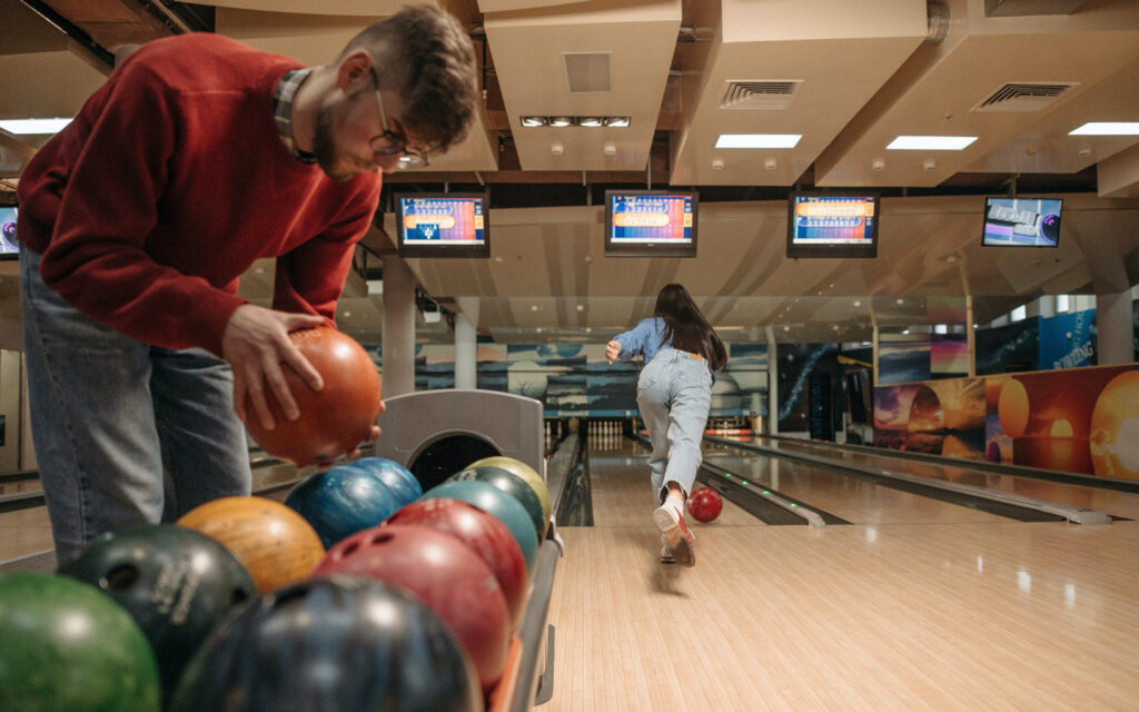 Ein Mann nimmt gerade eine Bowling-Kugel auf und im Hintergrund spielt eine Frau Bowling.