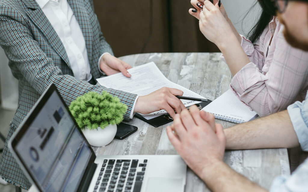 Drei Personen sitzen an einem Tisch mit Unterlagen und einem Laptop,