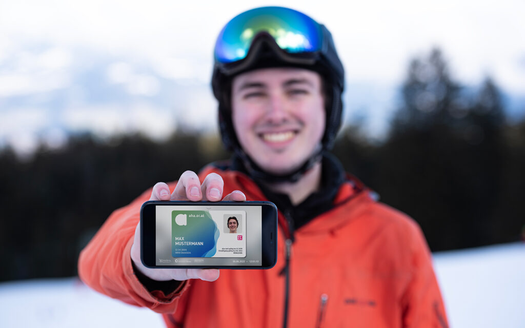 Junge in Skikleidung und auf der Piste stehend h&auml;lt die aha App auf dem Smartphone in die Kamera